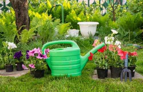 A garden scene featuring a neatly maintained lawn with vibrant green grass, bordered by several potted flowering plants with pink, white, and purple blooms. In the background, there are lush green shrubs, small trees, and dense foliage, creating a natural outdoor environment typical of a residential garden in Aldgate. A green plastic watering can with a red spout is placed on the lawn, alongside a small gardening fork and a potted plant, indicating active gardening care. The area appears to be well-lit under natural daylight, suggesting a clear weather condition. The garden layout includes a mix of plant containers and ground-level planting beds, providing a tidy and organized outdoor space suitable for professional gardening services such as hedge trimming or lawn maintenance. Overall, the scene reflects a healthy, well-kept garden environment that a gardening service like Hedge Trimming Aldgate would support, with a focus on flower beds, lawn, and general outdoor upkeep.