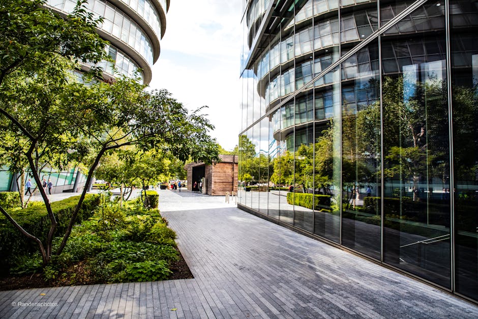 A modern urban garden situated alongside a sleek, reflective glass building with curved architecture in Aldgate. The garden features a variety of green plants, including a small tree with slender bran